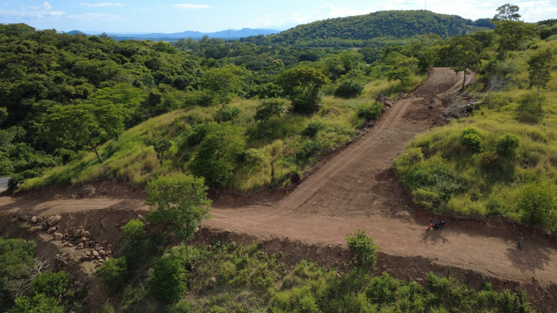 Vista aérea del proyecto El Paraíso en Chalatenango, con acceso de tierra rodeado de vegetación y montañas al fondo